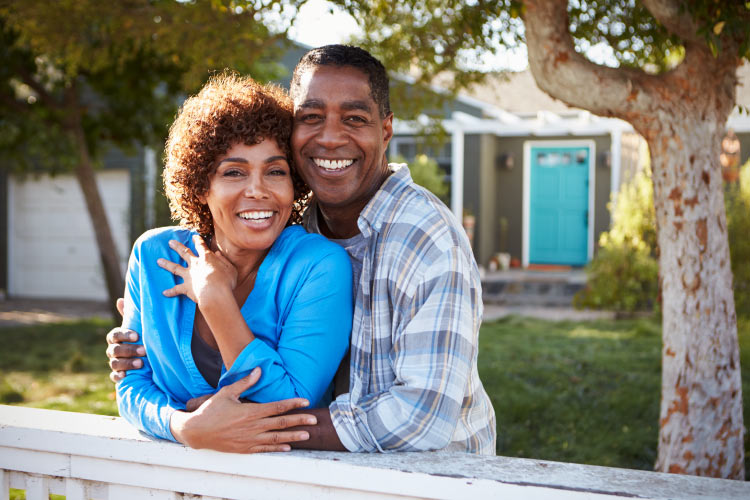 Brunette husband and wife embraces while smiling with their dental implants in front of the home with a blue door